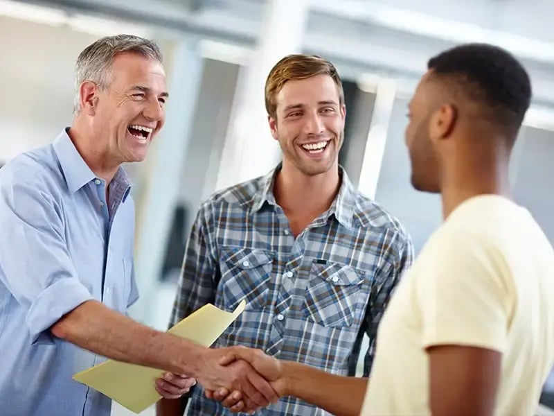 Two men shaking hands while a third man smiles during a friendly workplace conversation.